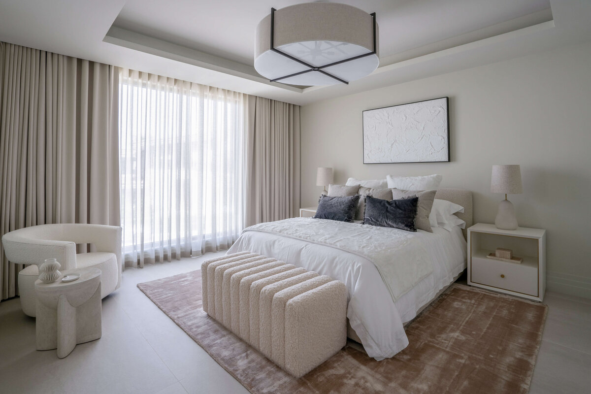 Light and airy guest room featuring a white bed, cream colored bench, and white side tables atop a soft tan rug. Above the bed is a white art piece. In the left corner, a contemporary white armchair. On the ceiling there is a large, round light. Floor-to-ceiling drapes cover large windows extending the length of one of the walls.