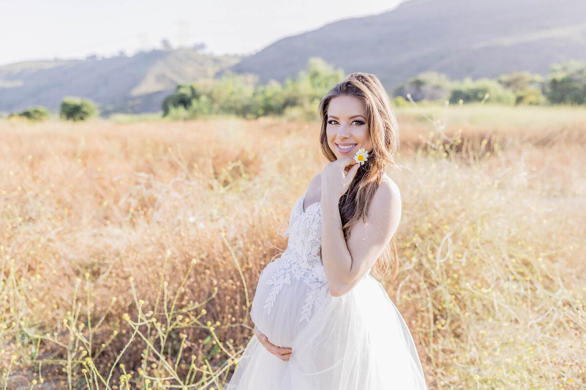 A pregnant woman in a white dress stands in a sunlit field, smiling and holding a small flower. She gently cradles her belly with one hand. Hills and trees are visible in the background.