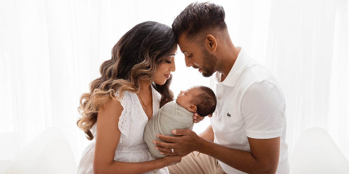 Mother and father sitting closely together, gently holding their newborn baby wrapped in light grey, with soft natural window light behind them.
