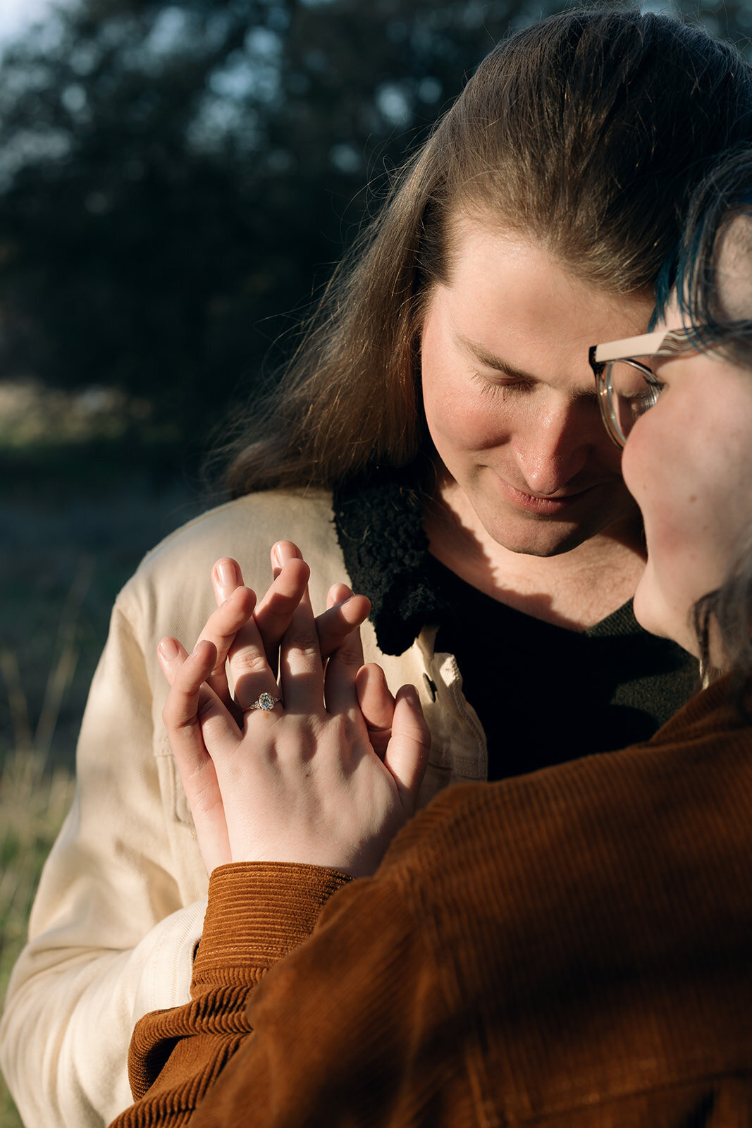 Romantic documentary-style photo capturing a tender moment between an engaged couple holding hands, showcasing her engagement ring.