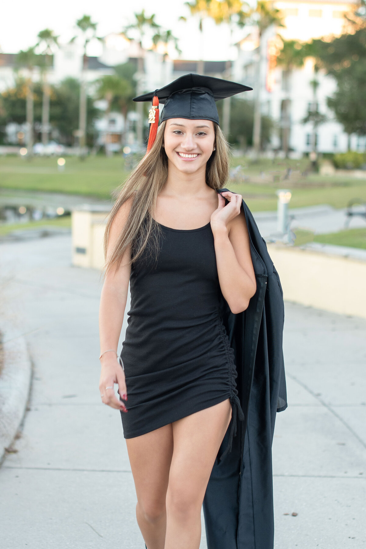 High school senior girl wearing cap walks holding gown over her shoulder.