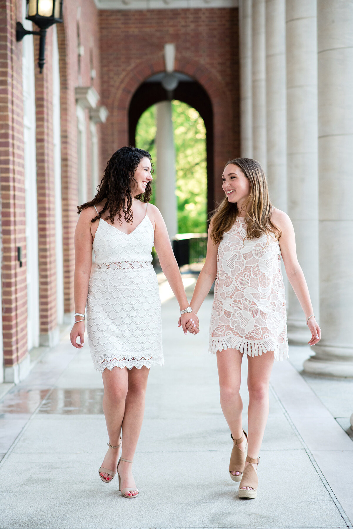 2 Best friends holding hands wearing cream mini dresses and walking together on the Vanderbilt campus near large pillars