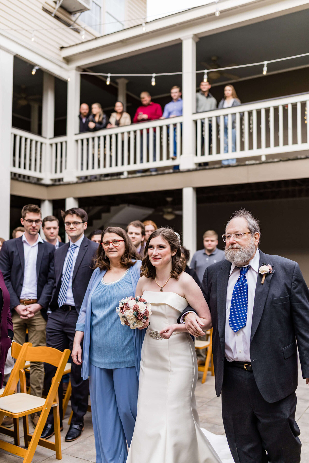 A bride in a white dress walks down the aisle, holding a bouquet and accompanied by her parents, as guests seated and standing watch the ceremony in an outdoor courtyard with string lights.