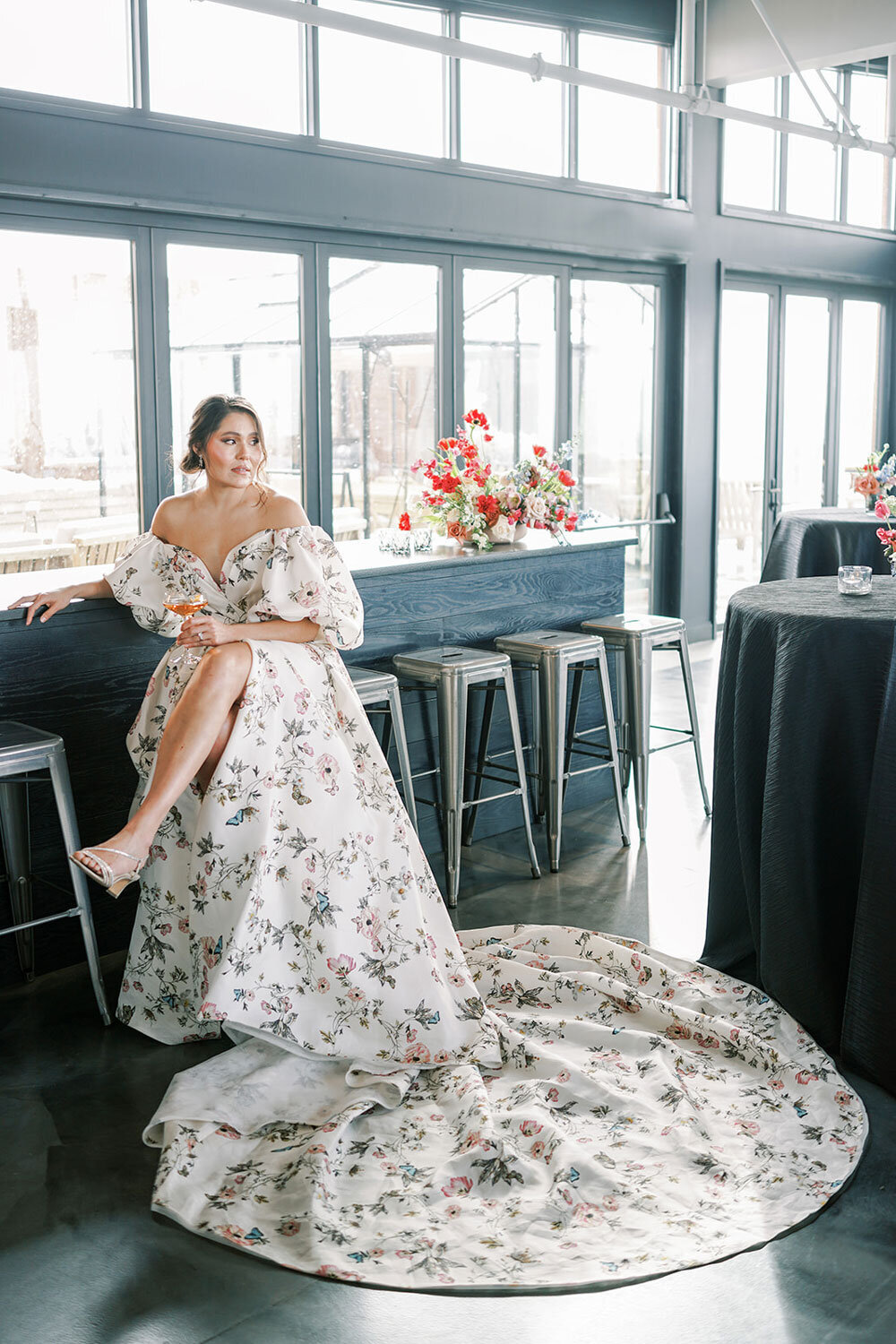 Bride wearing a floral off-the-shoulder gown with a dramatic train, seated at the rooftop bar of the Kimpton Armory Hotel in Bozeman, Montana.