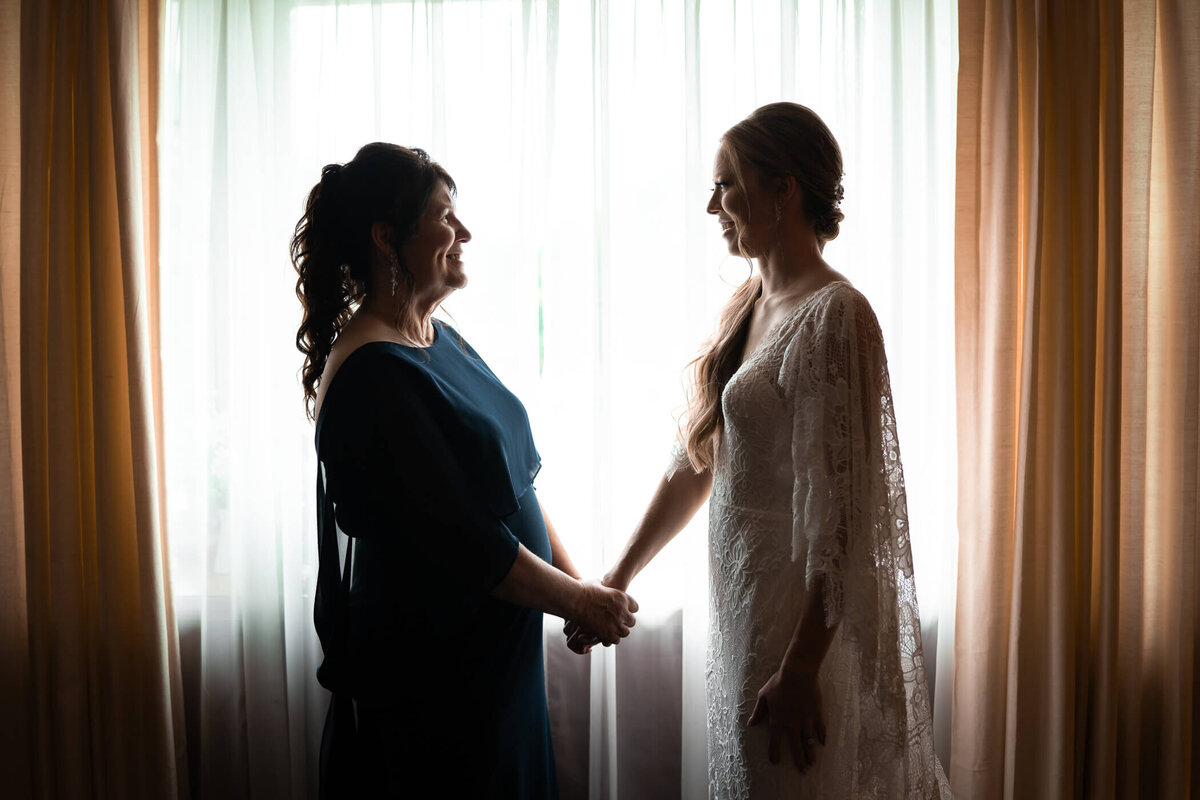Candid wedding photo of bride and her mother holding hands and smiling by window light at her childhood home in Pittsburgh, PA