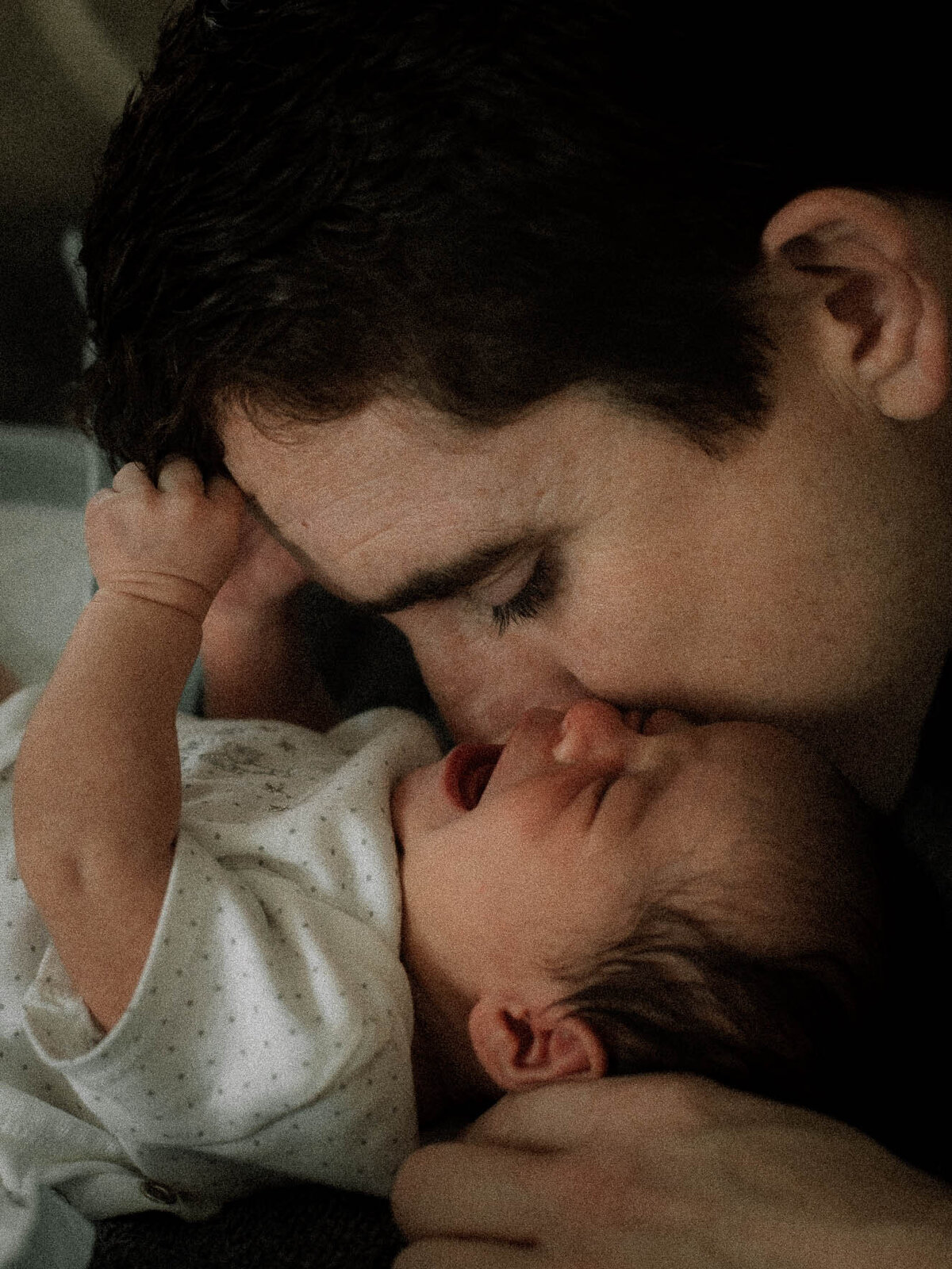 Father kissing newborn baby’s face while baby cries, emotional family photography moment.