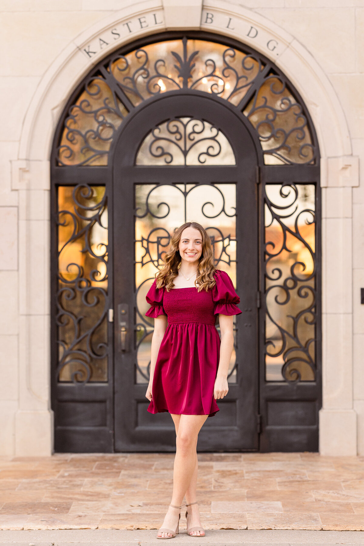 Smiling high school senior in a puff-sleeve red dress posing in front of an ornate black iron door at Adriatica Village in McKinney, Texas. The photo features golden-hour lighting and elegant European-inspired architecture.