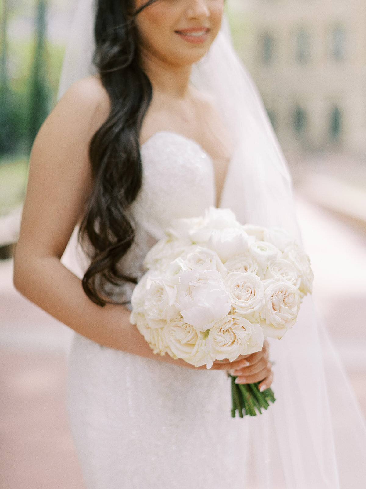 A bride in a strapless white wedding gown holds a bouquet of white roses and peonies at her classic Calgary wedding.