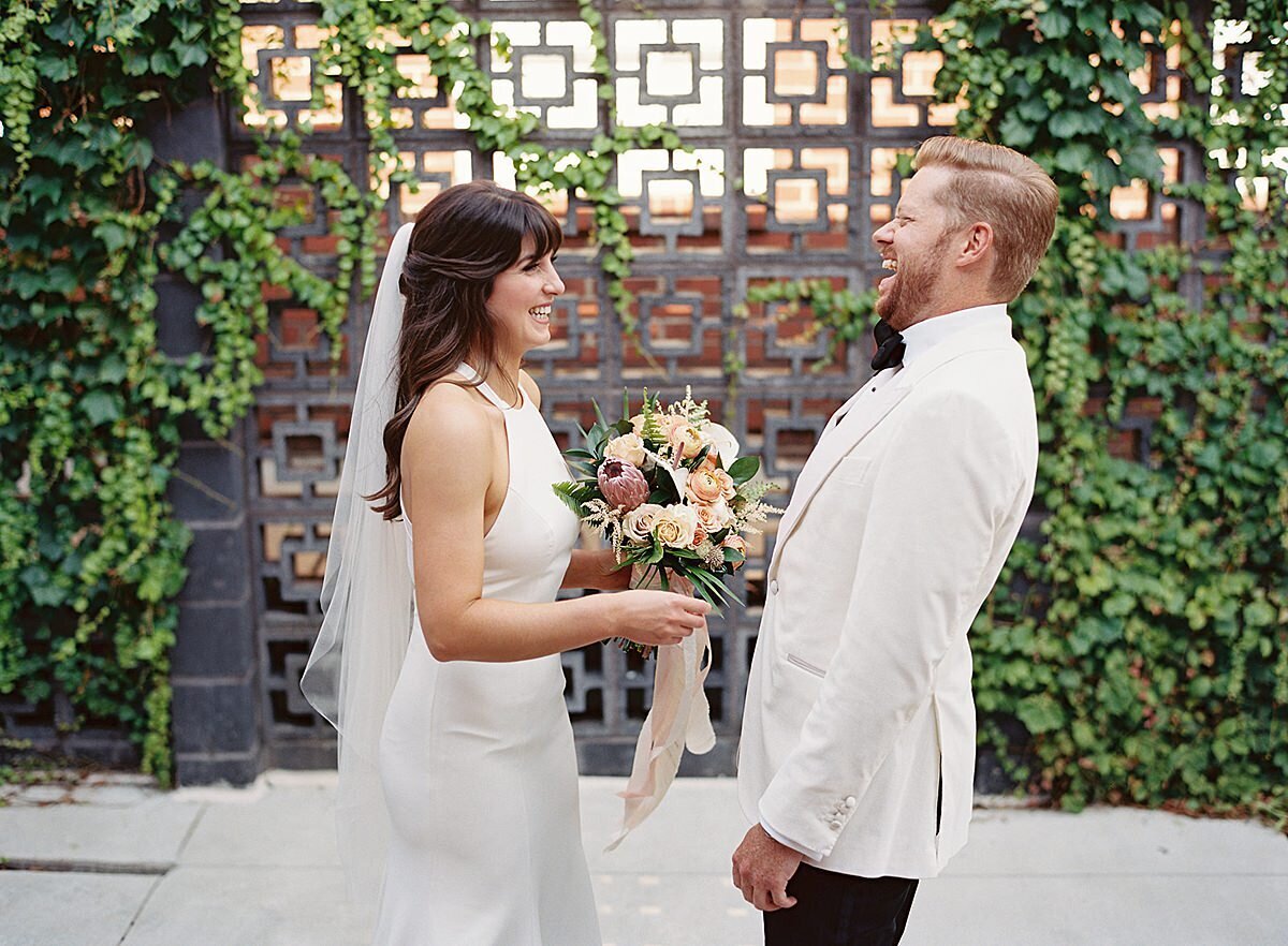 The bride wearing a white sheath dress and veil, holding a tropical bouquet of brightly colored flowers has a first look with the groom wearing a white jacket and black pants in the courtyard of Clementine Hall in Nashville