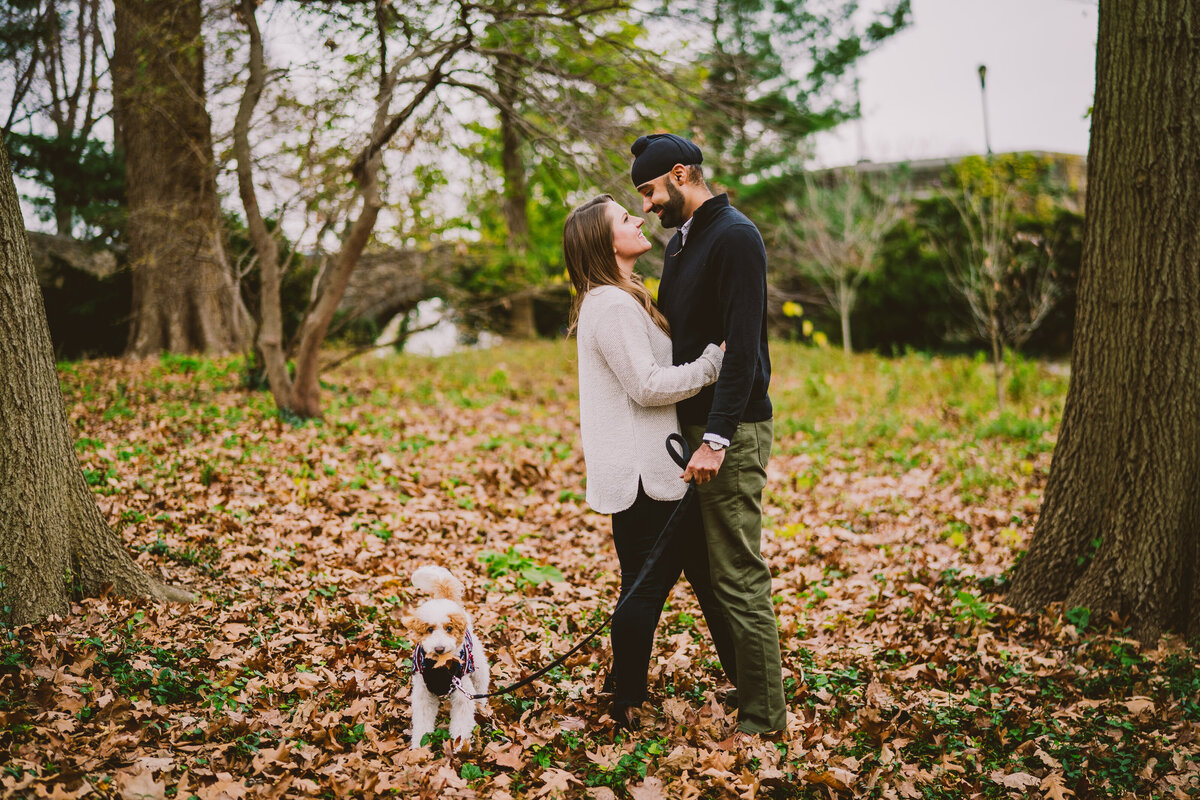 A couple sharing a moment standing on the fall leaves while their dog has a leaf in its mouth