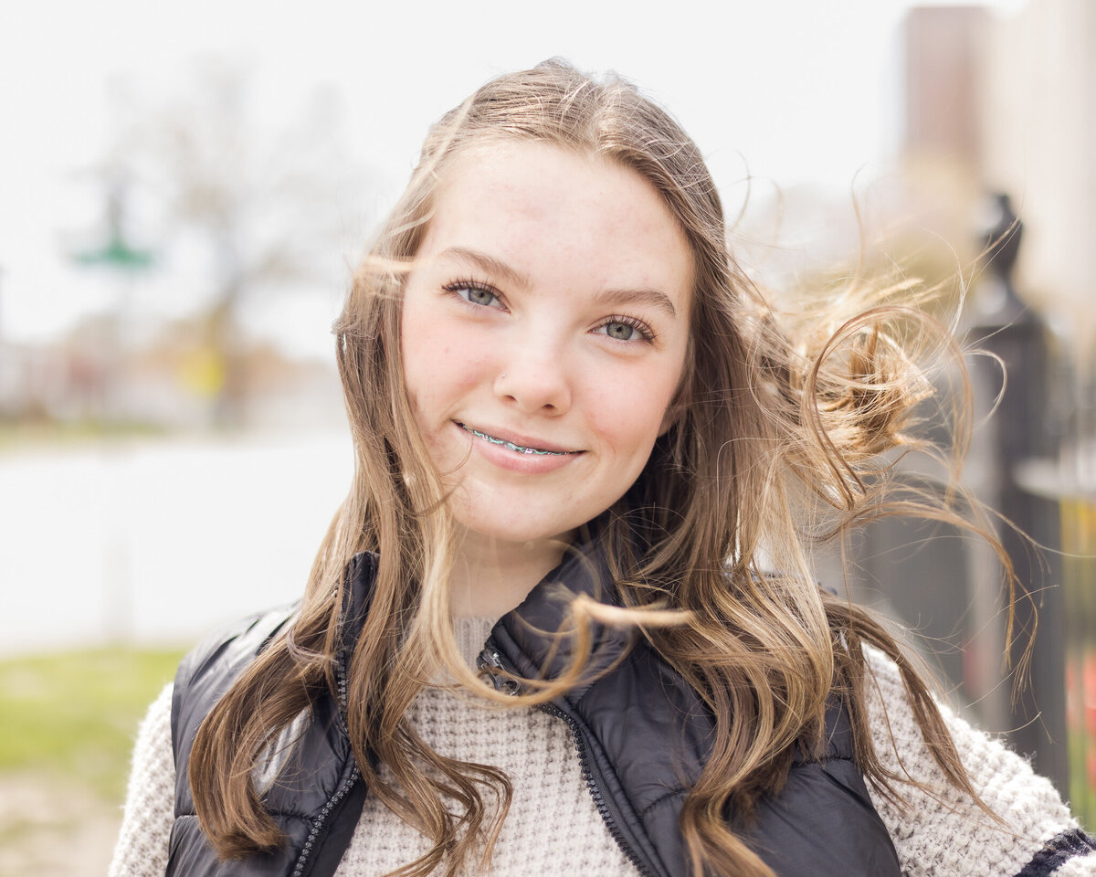 high school senior female, with long brown hair, wearing a black zip up jacket over a light tan long sleeve shirt, looking at the camera, smiling