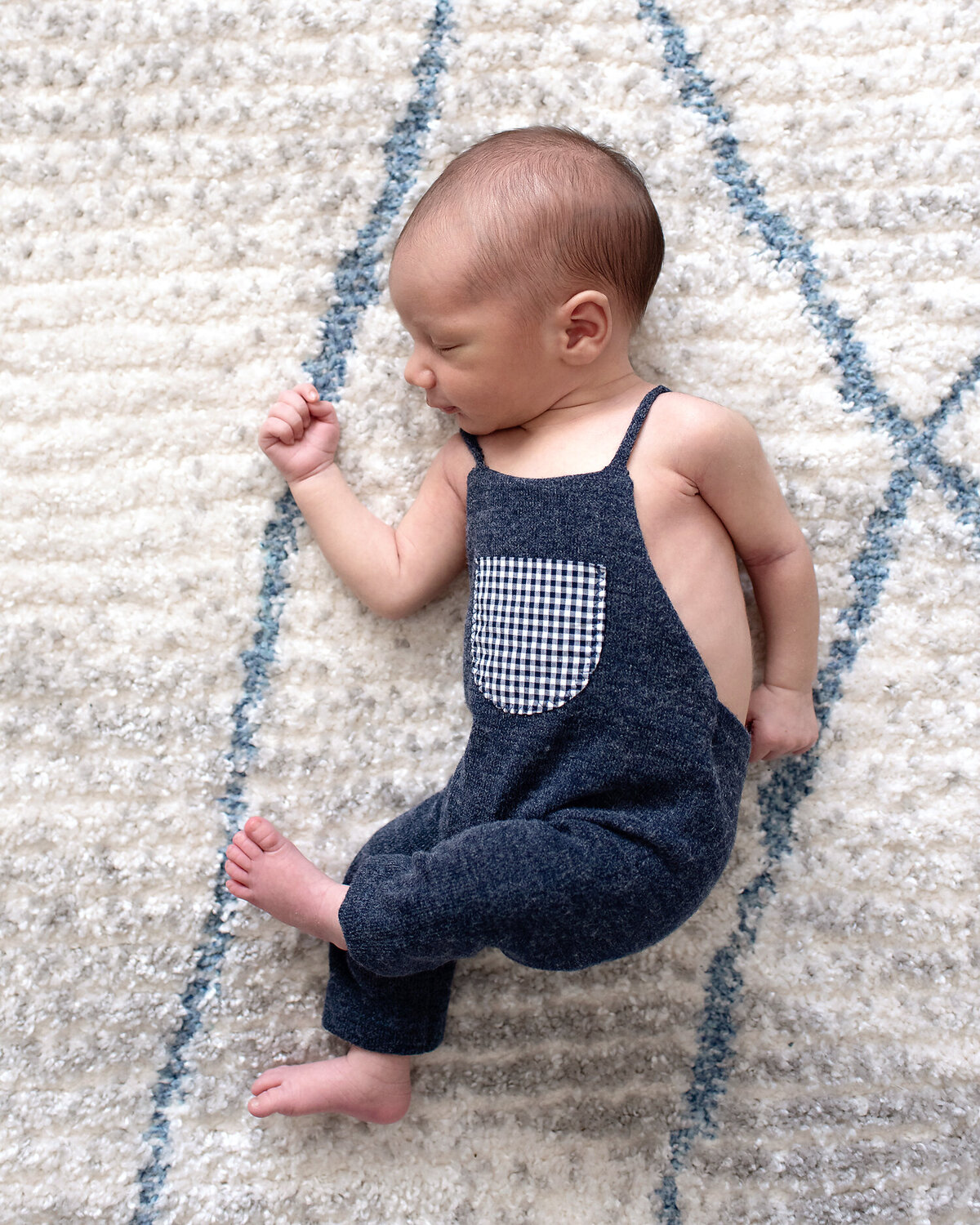 Newborn boy in a blue romper laying on a white and blue rug in his nursery.