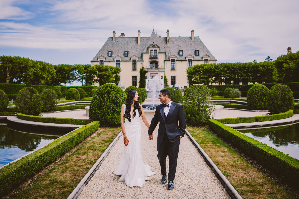 Bride and groom walking and looking at each other in a garden with Oheka Castle's fountain in the background
