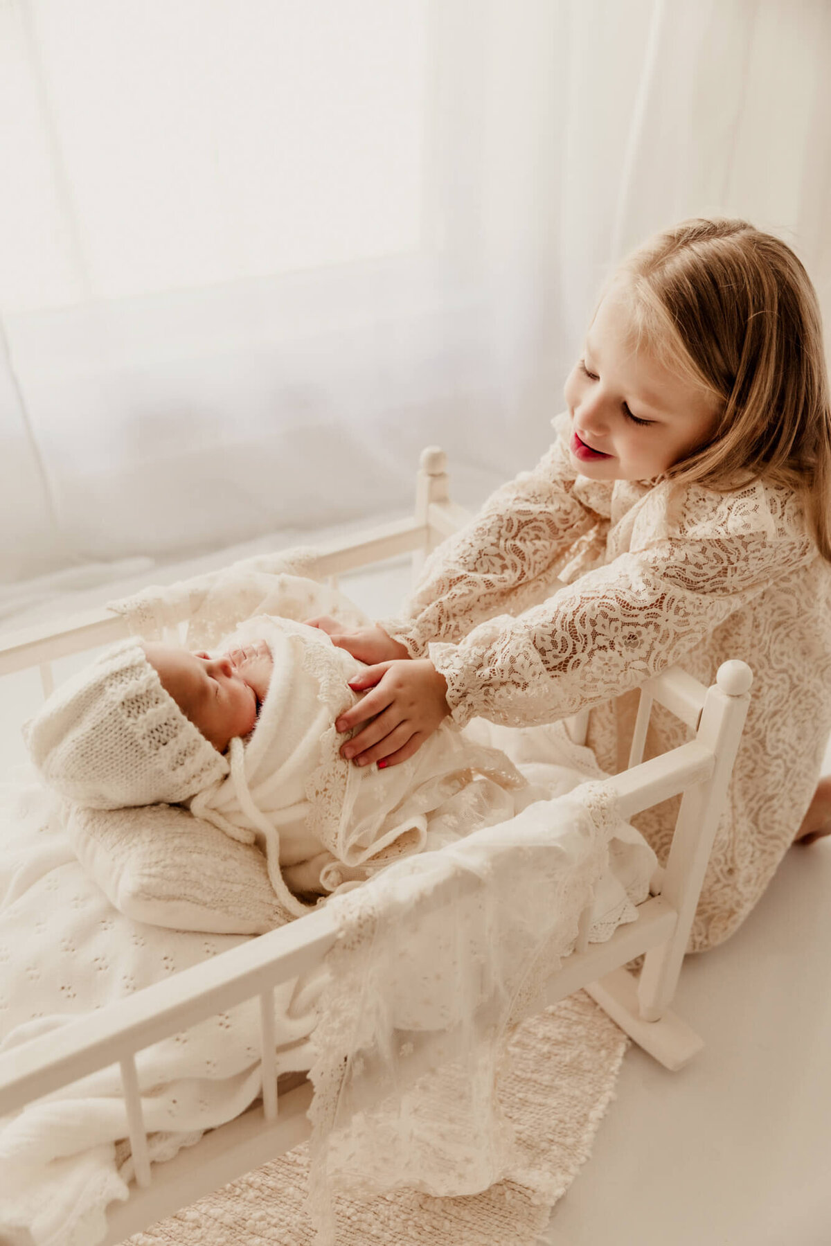 Older sister admires her new baby sister who is laying in a white cradle.