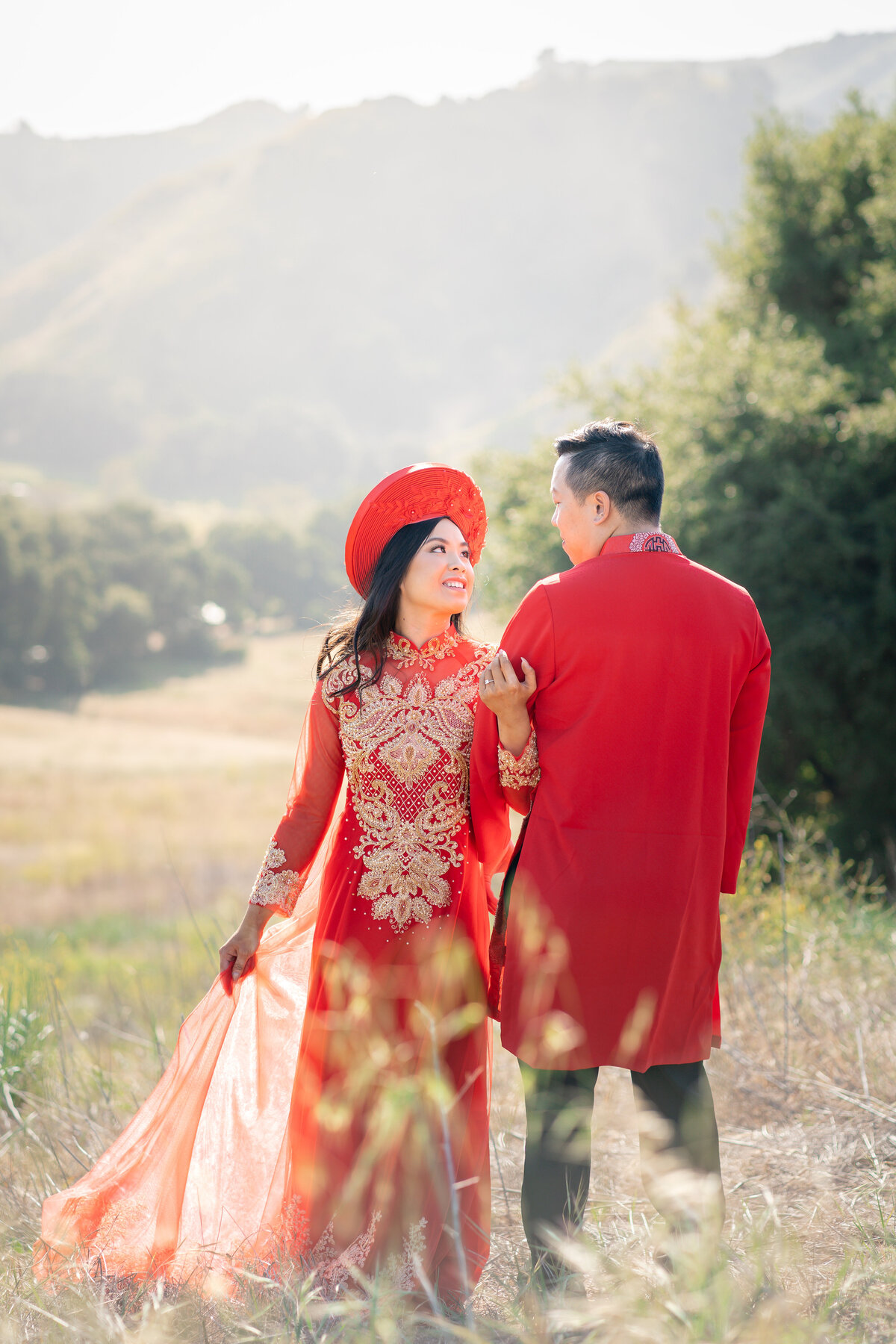 Asian bride and groom couple eloping at Malibu Sate Park wearing Vietnamese red Ao Dai red within golden glow of wildflower field and mountains.