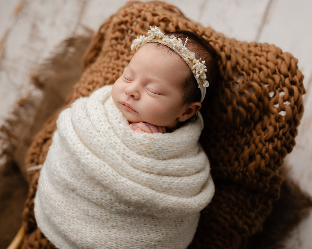 Peaceful newborn baby girl wrapped in soft ivory yarn, adorned with a delicate floral crown, resting comfortably on a cozy textured backdrop. Captured by Inland Empire newborn photographer Kaitlyn Dawn Photography.