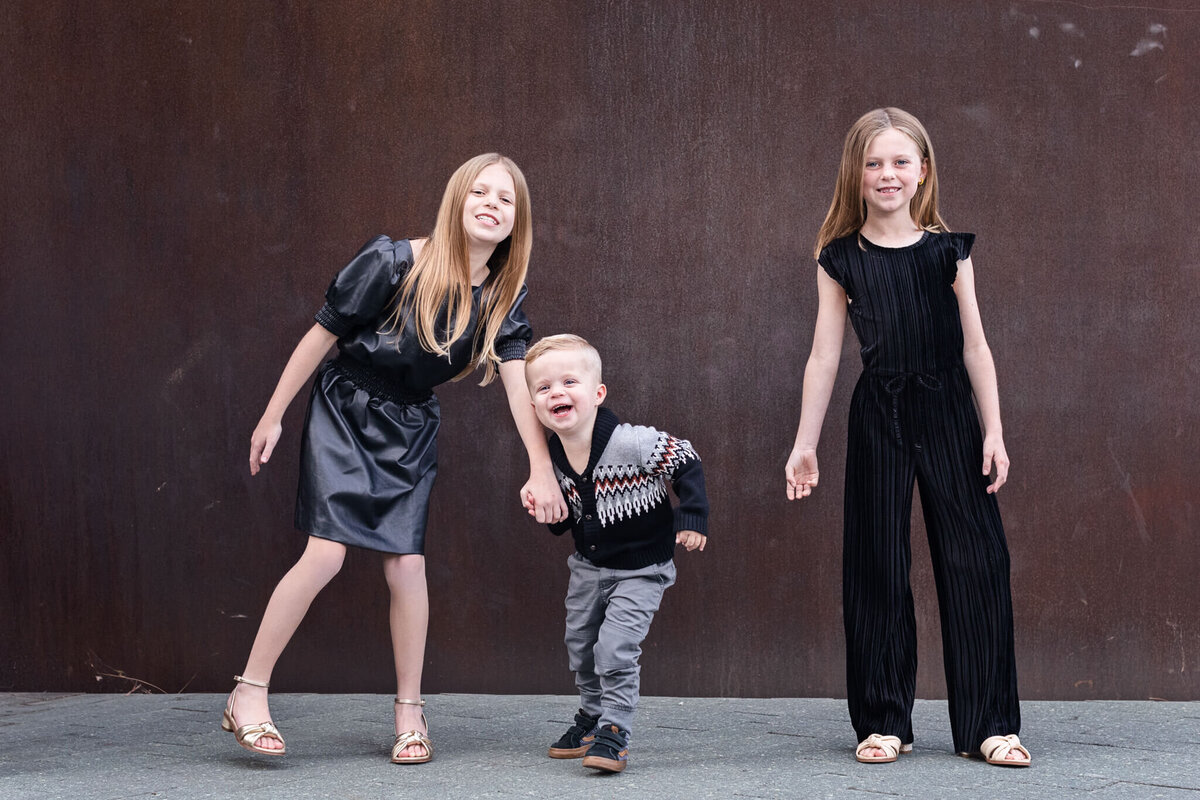 Two young sisters wearing black standing on either side of their little brother with blonde hair, laughing and looking at the camera in front of a dark brown wall. Captured at OCMA.