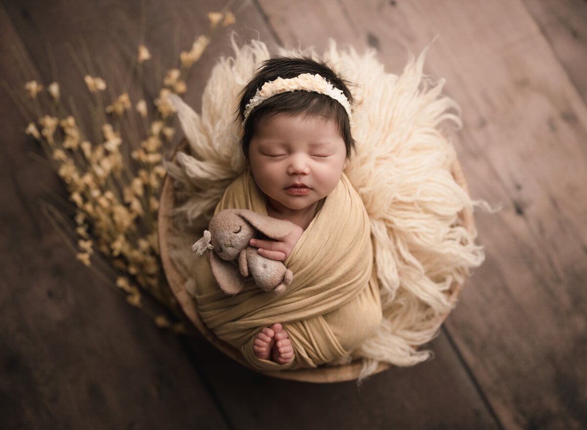 Aerial image of Riverside, CA newborn photoshoot. Baby girl is wrapped in a muted yellow swaddle with her fingers and toes peeking out. She is holding a small felt bunny. Captured by Riverside's best newborn photographer Bonny Lynn Photography.