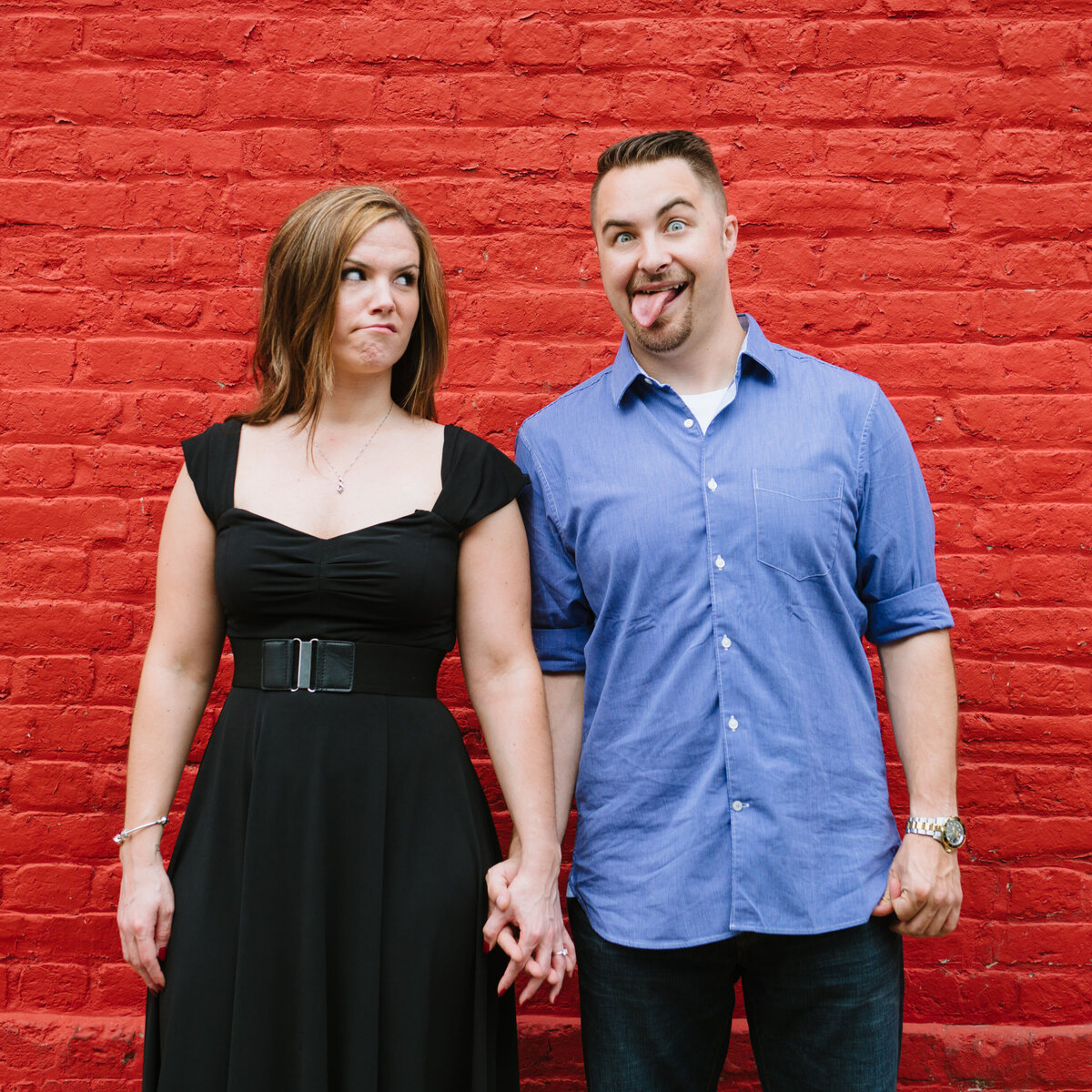 A couple holding hands and making funny faces while standing in front of a red wall.