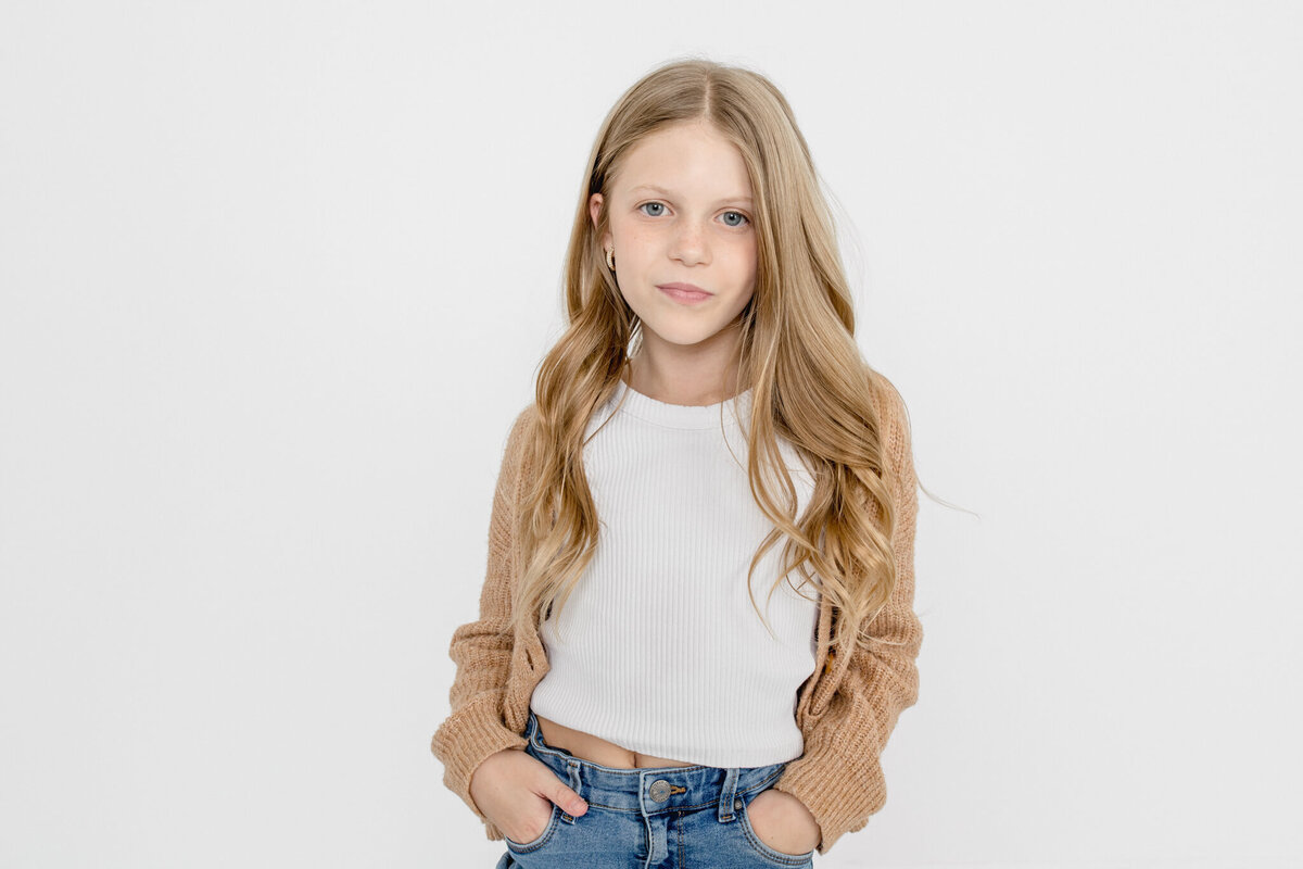 A young girl with long, blonde hair, wearing a white shirt, jeans, and light brown cardigan, putting her hands in her pockets and looking serious at the camera.