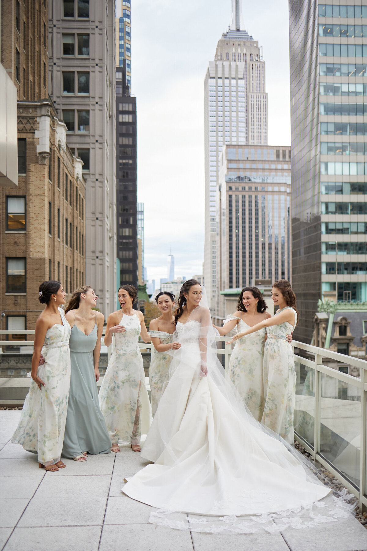 This is a stunning urban rooftop setting for a wedding photo, isn't it? The photo was taken in New York City, specifically at The New York Public Library by photographer Shawn Connell. The bride looks radiant in her long white gown, standing with her five bridesmaids who are elegantly dressed in light-colored dresses. The backdrop of the cityscape with tall buildings adds such a dramatic touch to the scene. Additionally, videographers Aaron Navak Films captured the beautiful moments of the wedding day.