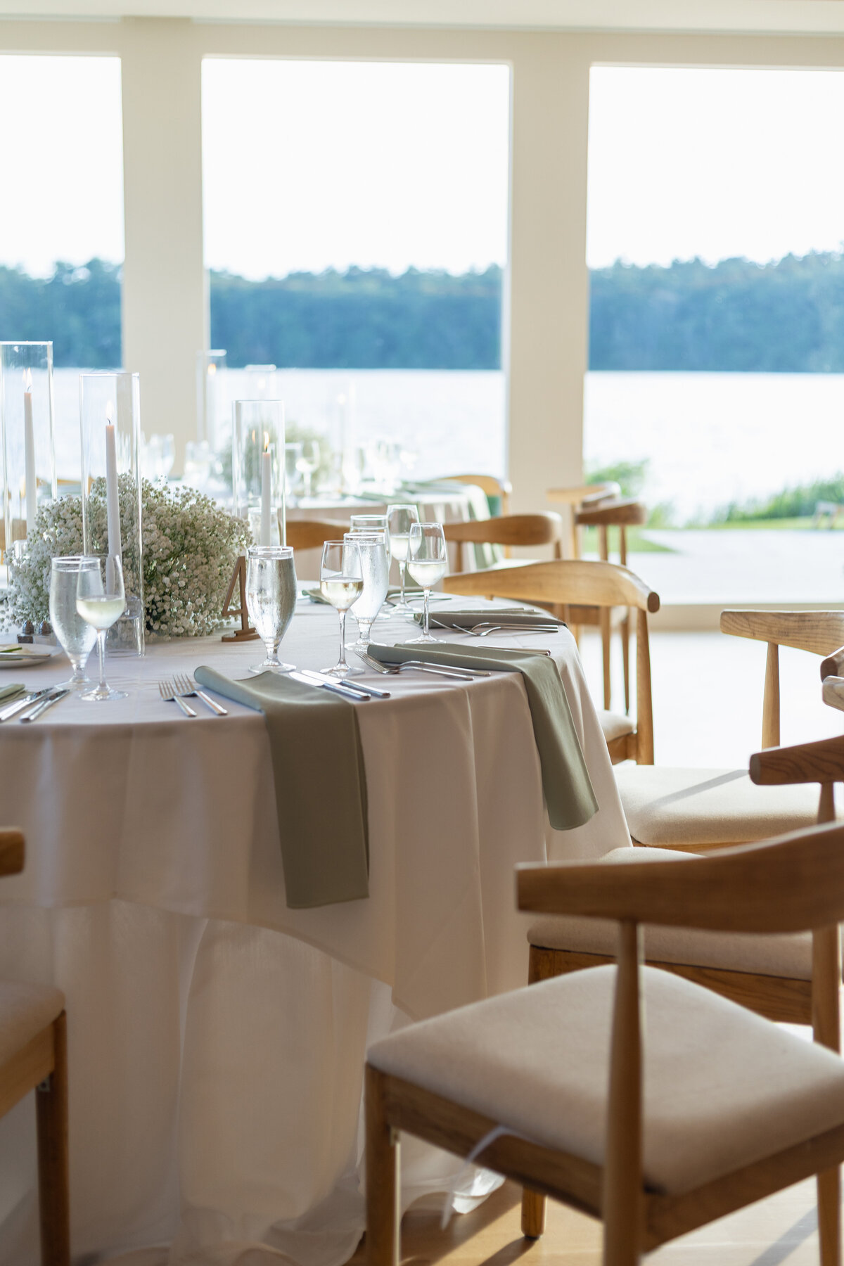 The Lakehouse Inn MA  | Kelsey Sheehan Photography Timeless Rhode Island Weddings | Elegant lakeside dining setup with a round table dressed in white linen, adorned with glassware and candles, overlooking serene water and lush greenery.