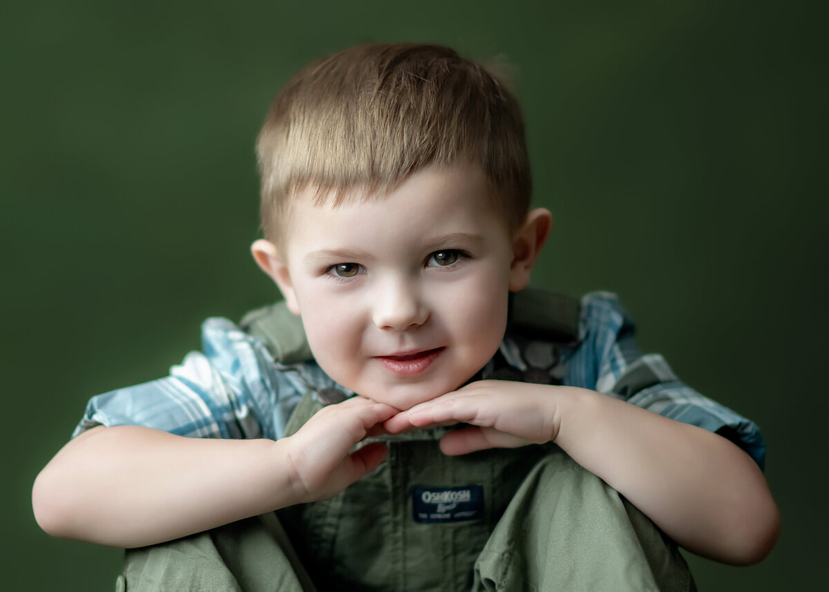 Child in plaid shirt and overalls with his hands under his chin on a green background.