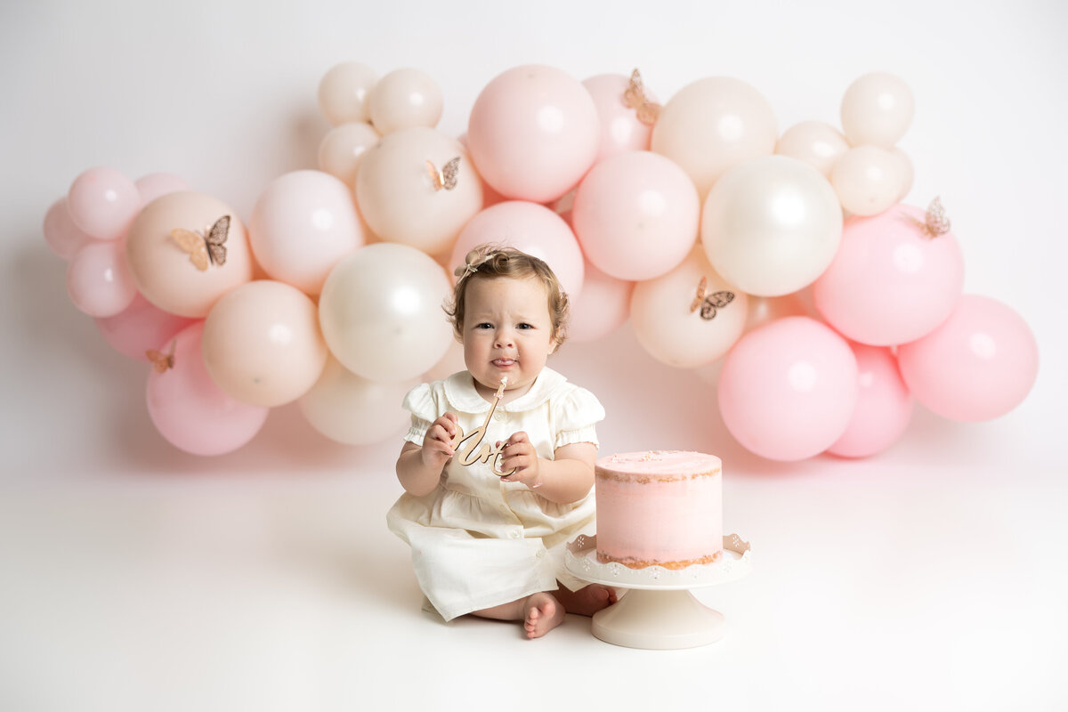 Baby girl cake smash with pink and white balloons, butterflies, and pink cake
