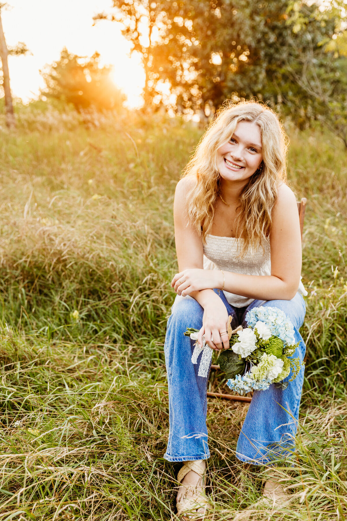 young blonde teen girl in a white top and jeans sitting on a wooden chair in a field at sunset while holding a bouquet in a park in Neenah.