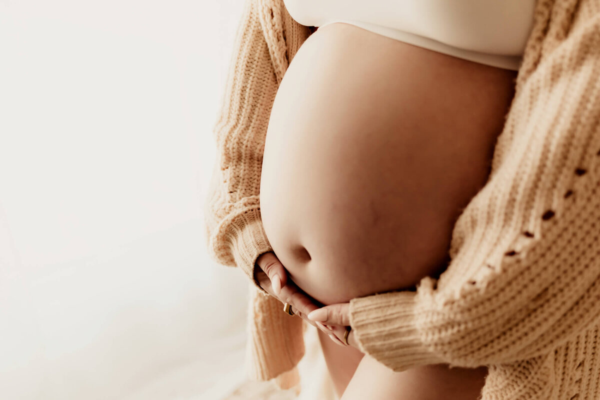 Woman holding her pregnant belly with a beige long sleeve sweater. 