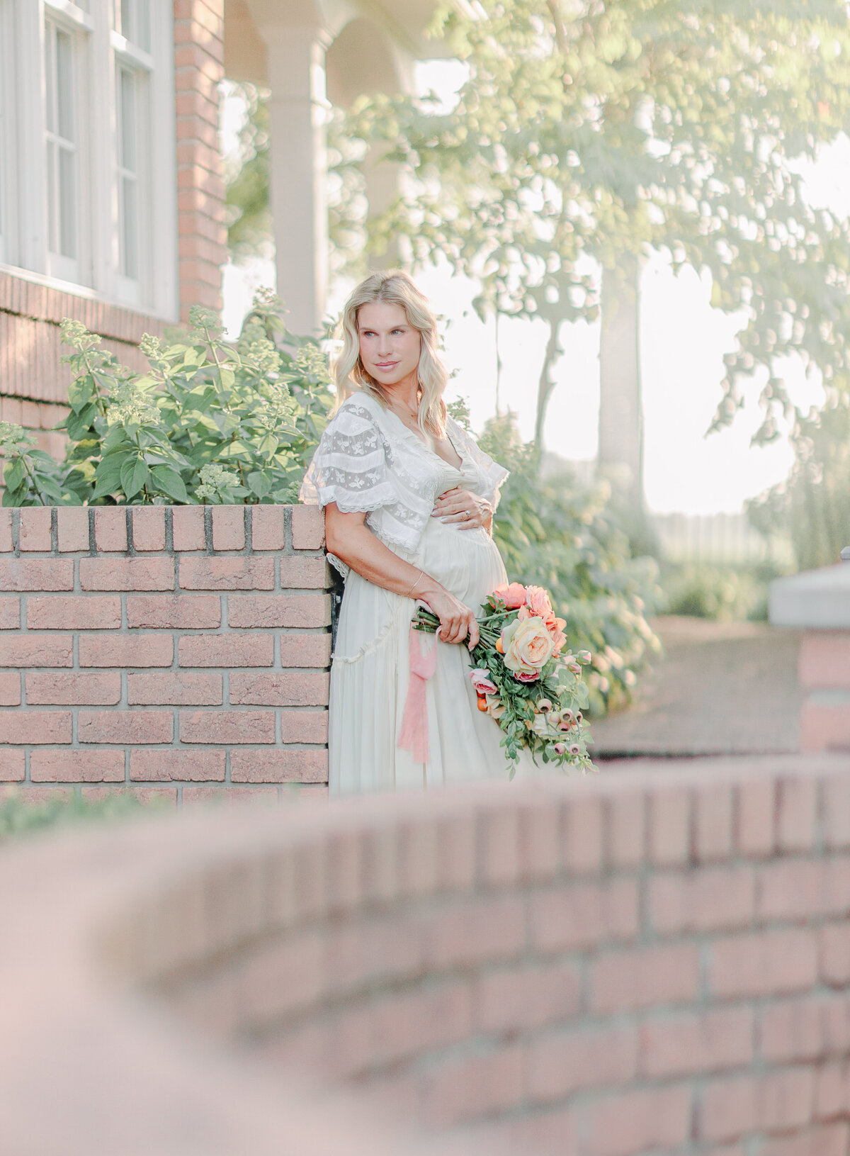 Pregnant woman in a flowing white dress holding a bouquet of flowers during a golden hour maternity session, photographed outdoors in Eau Claire, Wisconsin. This soft, light-filled image captures the beauty and emotion of motherhood photography with a natural, timeless style.