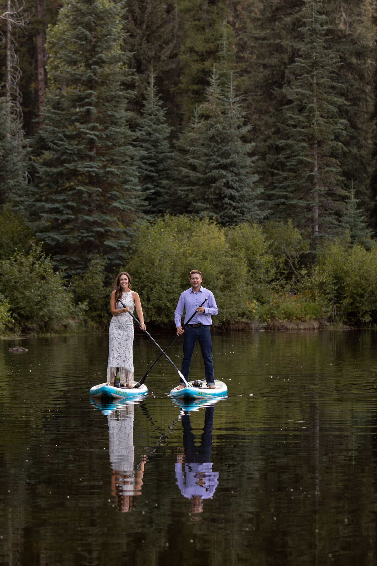 A bride and groom stand on paddle boards in the middle of a lake with their paddles crossed across each other's board.