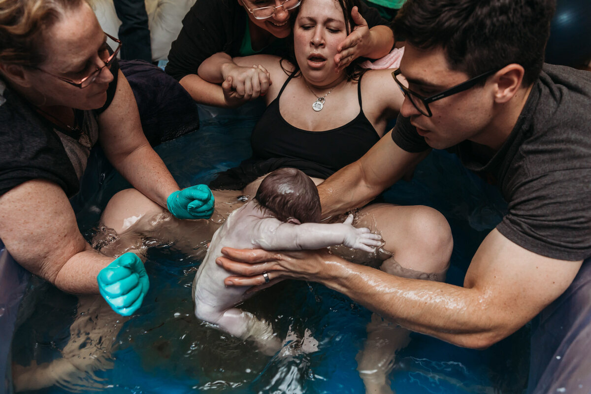 woman in birth pool as man pulls baby out of birth water