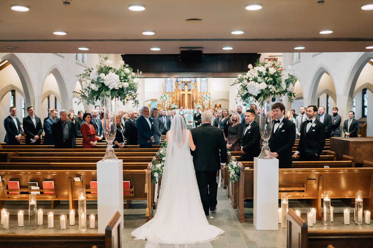 Black and white photo of a bride wearing a strapless dress with a bow and groom with a black tie tux walk hand in hand. Photo by Anna Brace, an Omaha wedding photographer.