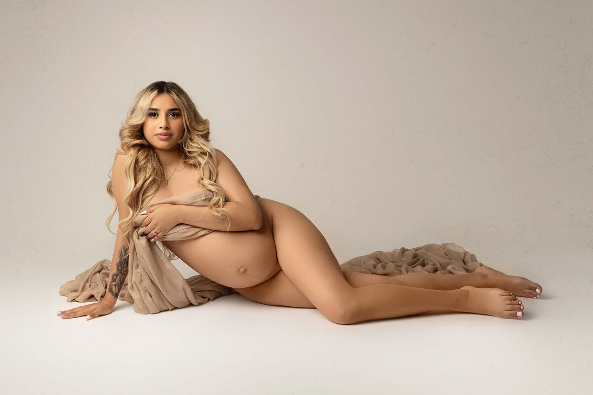 An expecting blonde woman wraps her chest in a tan sheet while laying across a studio floor taken by New Orleans Maternity Photographer Melinda Gilmore