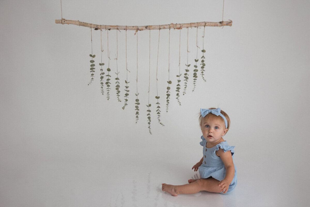 A baby girl in a blue romper and matching headband sits on a white floor, looking up. Above her, greenery hangs from strings attached to a wooden branch.