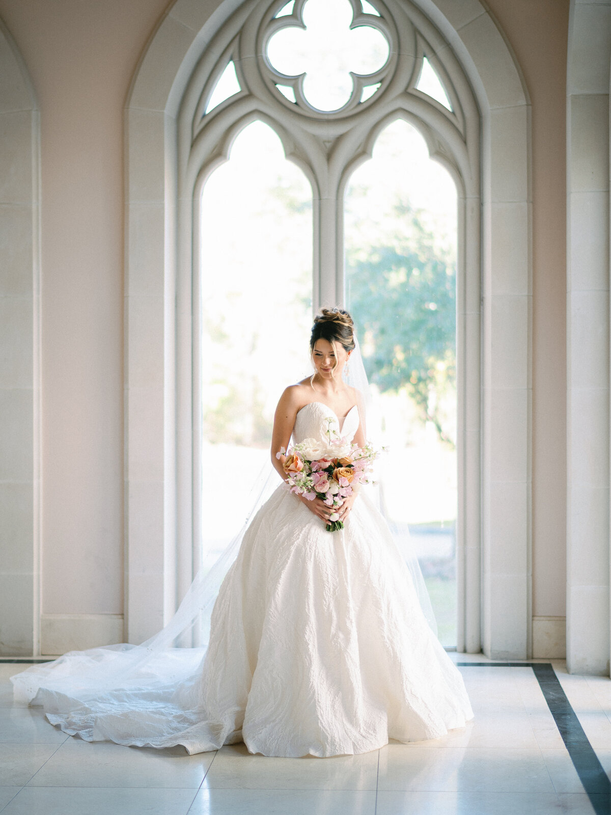 Bride by window in beautiful gown smiling with pretty bouquet