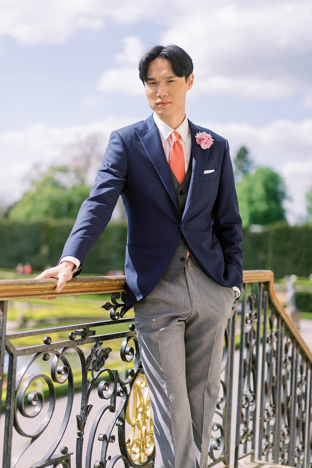 groom wearing a navy suit jacket and grey trousers with a coral tie standing on stairs in the garden of luxury wedding venue blenheim palace
