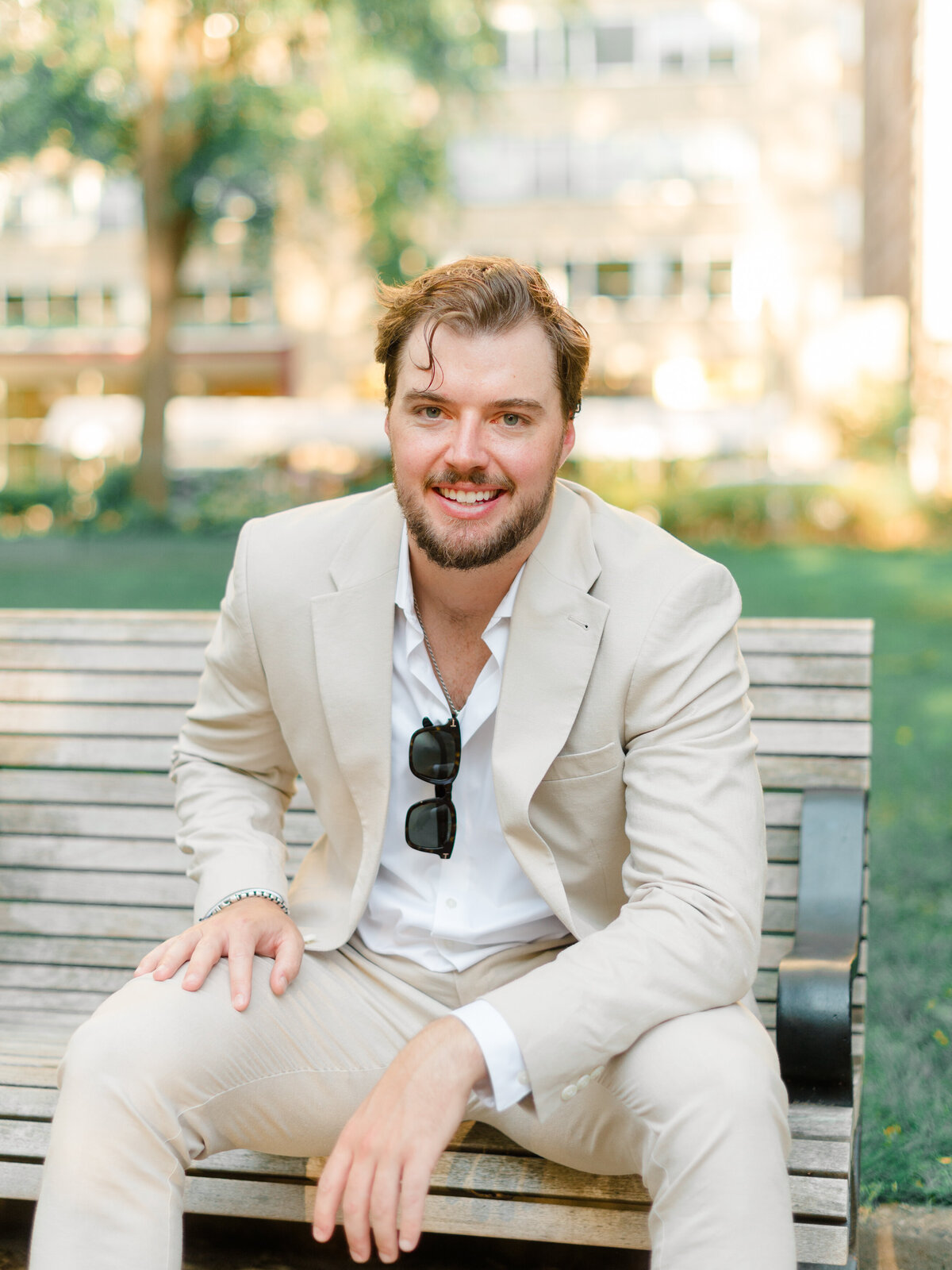 A groom-to-be sits casually on a park bench in Rittenhouse Square, wearing a light beige suit and white shirt with sunglasses tucked into his jacket. His relaxed smile and confident presence are captured in soft, golden light by Elizabeth Kane Photography, a Luxury Philadelphia Wedding Photographer.