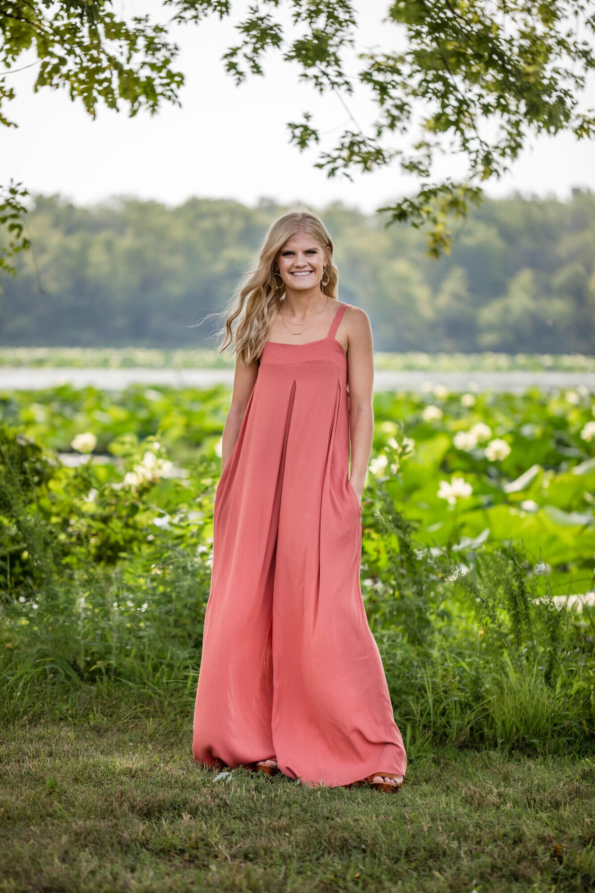 A senior picture of a lovely blonde teen in a coral summer jumper standing in front of a lake covered in waterlilies. Captured by Springfield, MO senior photographer Dynae Levingston.