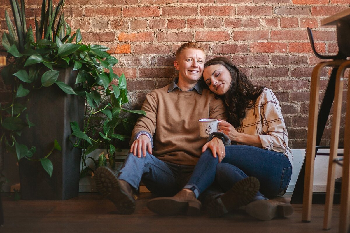 couple-sitting-against-brick-wall-in-coffee-shop-embracing-during-warm-moment-in-franklin-indiana-couples-photography-session