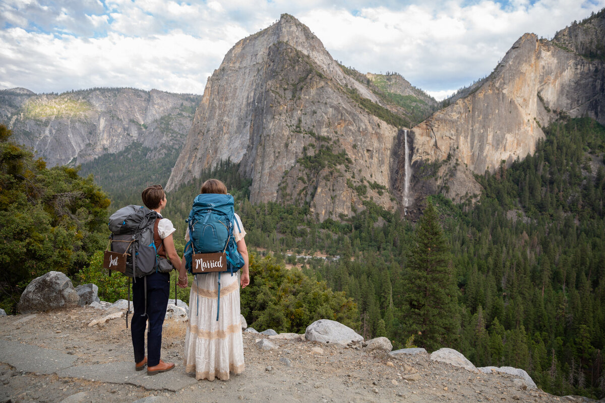 Two brides wearing backpacking packs hold hands and stare at a giant waterfall across the valley in Yosemite.