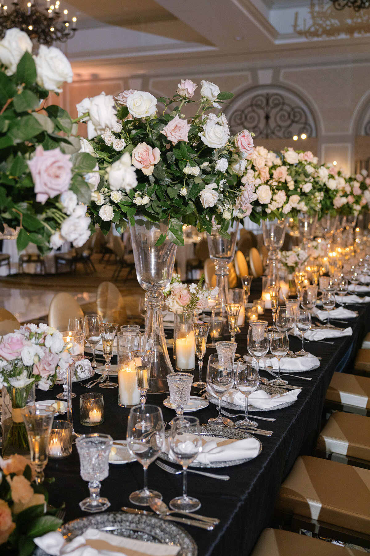 elegantly styled wedding reception table at The Adolphus in Dallas featuring a black tablecloth, tall glass vases with pink rose arrangements, numerous candles, and refined glassware and chargers.