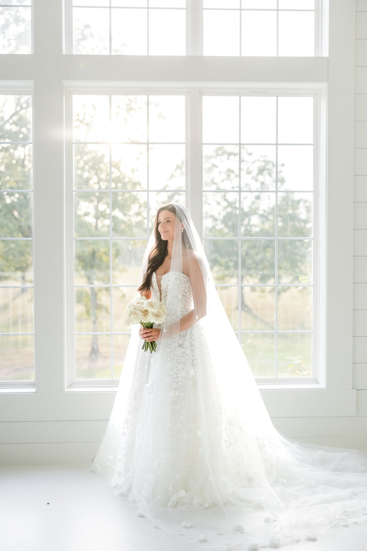 A bride smiles while standing under large windows for an Arkansas wedding photographer