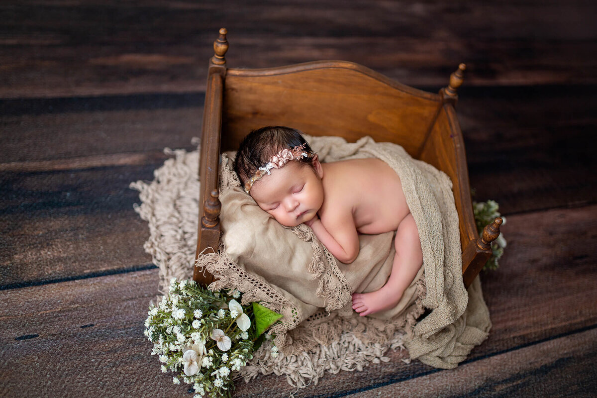 Classic boho newborn baby girl laying naked on a wooden daybed surrounded by beige flowers on a barnwood floor 