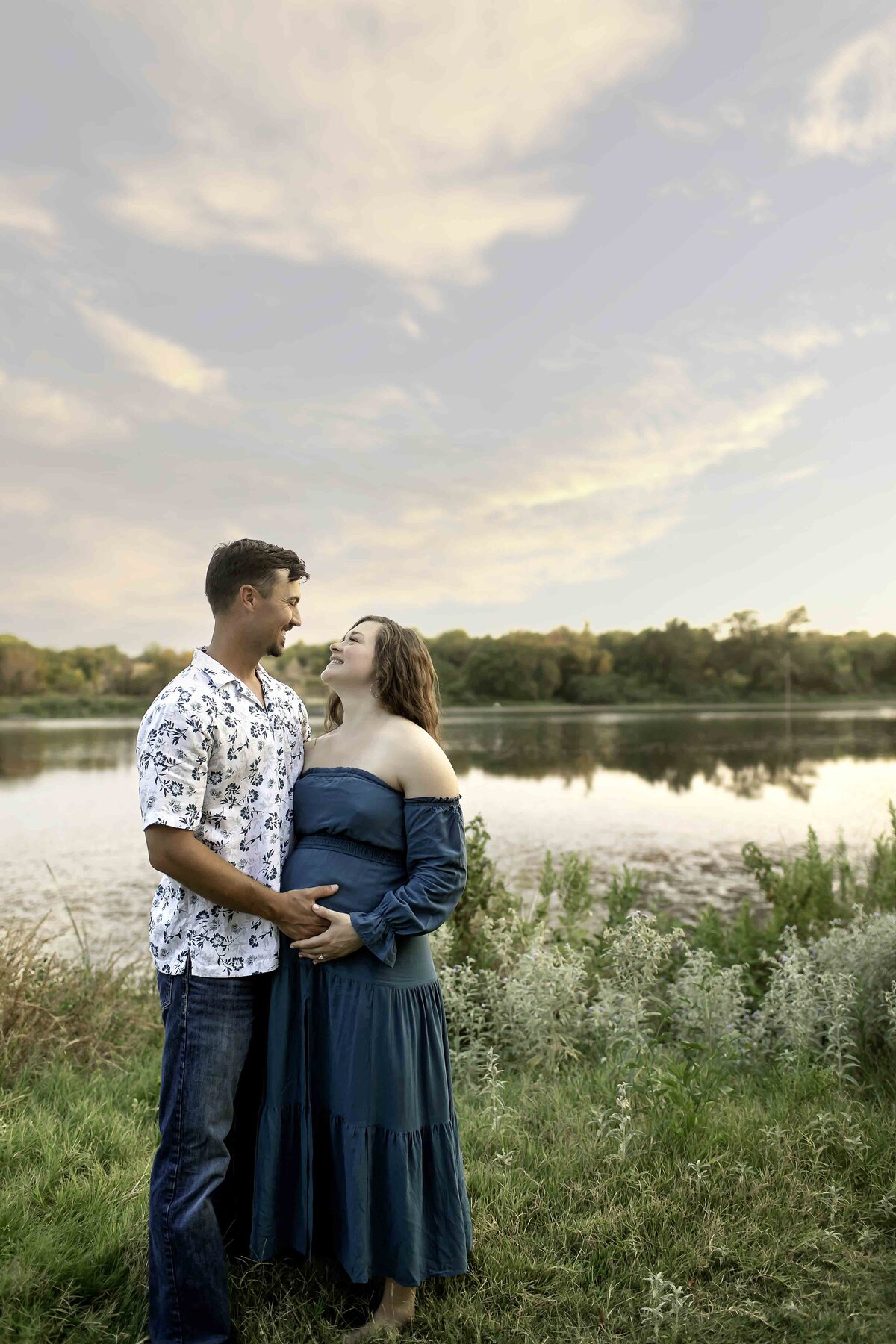 A romantic maternity photo of the expectant mom and dad standing by the lake, surrounded by lush greenery, with the dad gently resting his hand on his wife’s belly, captured in a tender moment by Chunky Monkey Photography.