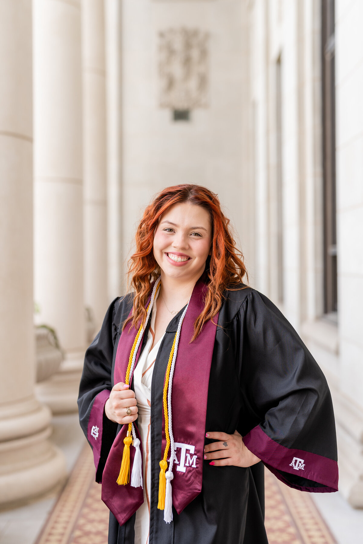 Texas A&M senior girl wearing grad gown, Aggie stole and cords with hand on hip while smiling in the columns of the Administration Building