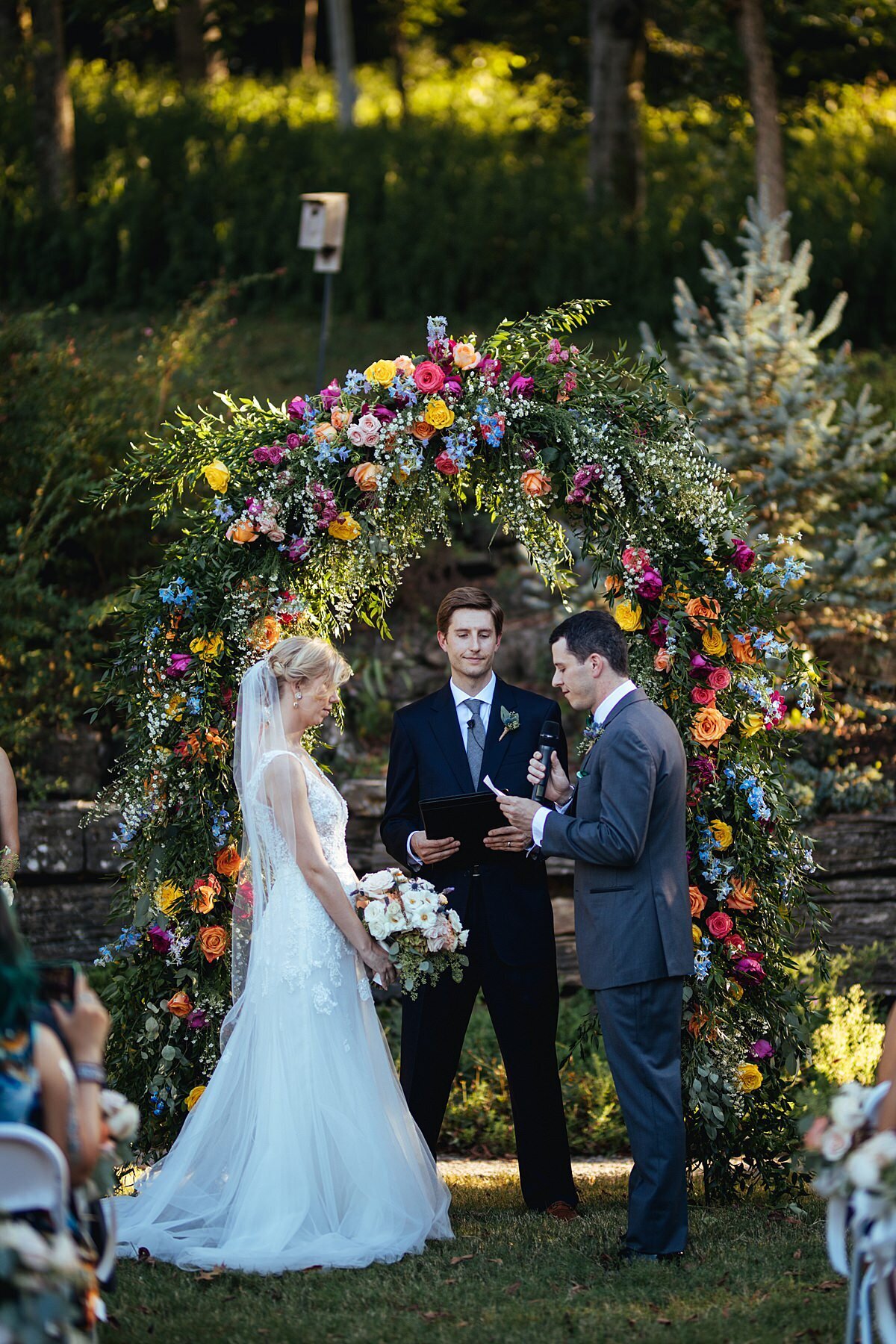 The bride wearing a long sleeveless lave wedding gown and a long sheer veil holds a large bouquet of white, ivory, blush and peach roses as the groom wearing a charcoal gray tuxedo holds a microphone and a piece of paper as he recites his vows to the bride. The wedding officiant weaing a black suit and grey tie stands behind them under the lush floral arbor. The floral arbor is covered in greenery and accented by red, hot pink, yellow, orange, blue and purple flowers for the wedding at Cheekwood.