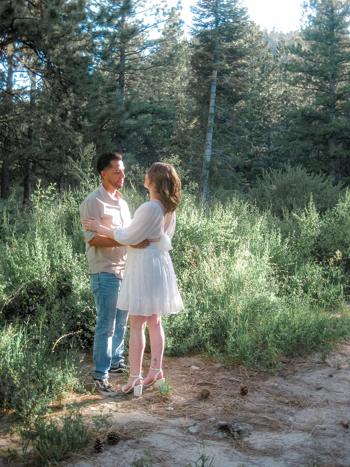 The couple stands closely together in a sunlit forest clearing, with the man in casual attire and the woman in a flowy white dress, surrounded by lush greenery.