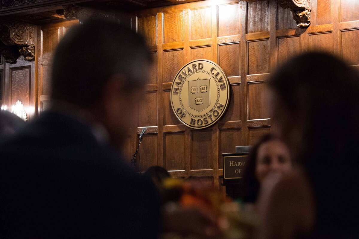 Harvard Club seal on wooden wall shot through crowd of guests in attendance for honored guest dinner